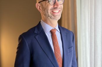 White Male with glasses and suit, standing and smiling